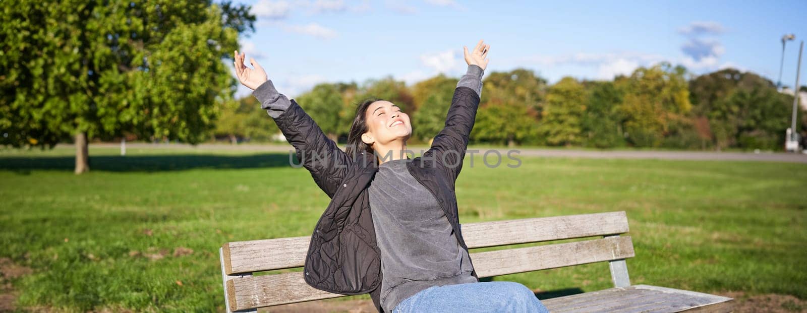 Happy asian woman stretching her hands, sitting on bench with excited face, smiling pleased, feeling freedom, enjoying day in park by Benzoix