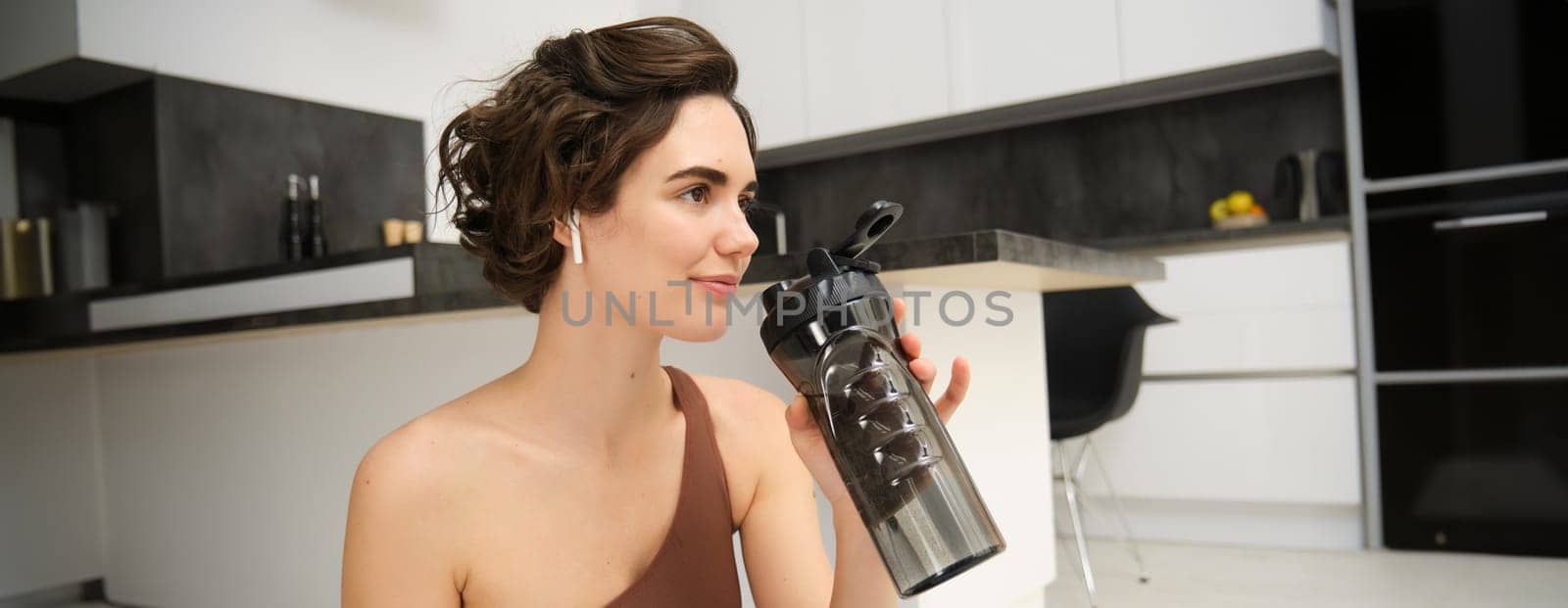 Portrait of fitness woman, young sportswoman at home, drinks water from bottle after workout, training exercises, takes a break after pilates, yoga training, stays hydrated.