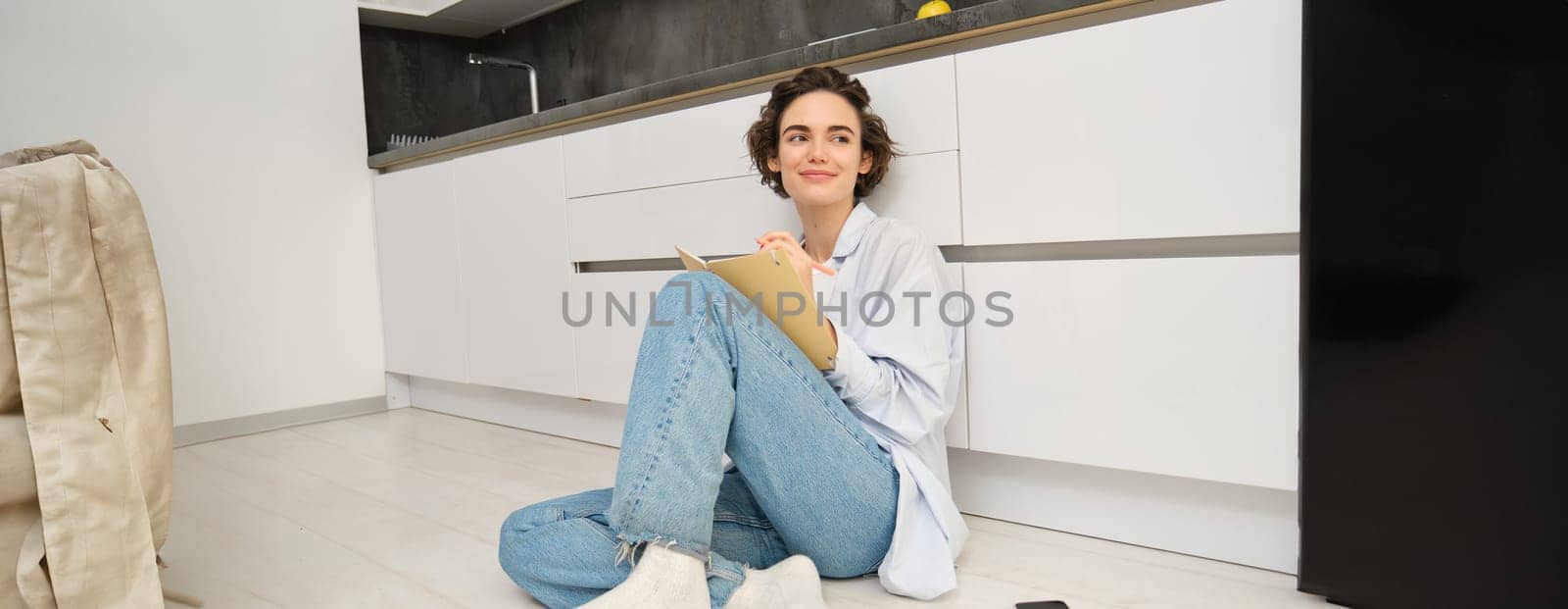 Portrait of cute brunette woman sits on floor, draws in her planner, writes diary on floor in kitchen and smiles. Leisure, lifestyle and people concept