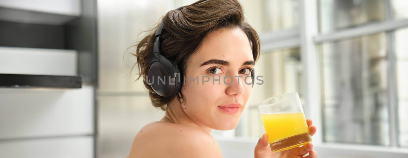 Sport and healthy lifestyle. Portrait of happy sporty girl, fitness woman in headphones, rewinds at home, drinks orange juice with happy smile after workout.