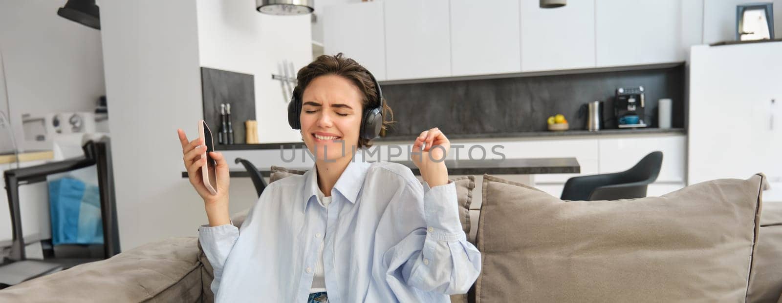 Portrait of beautiful, happy young woman, listening music in headphones, holding smartphone and dancing on sofa at home, enjoys spending time indoors alone.