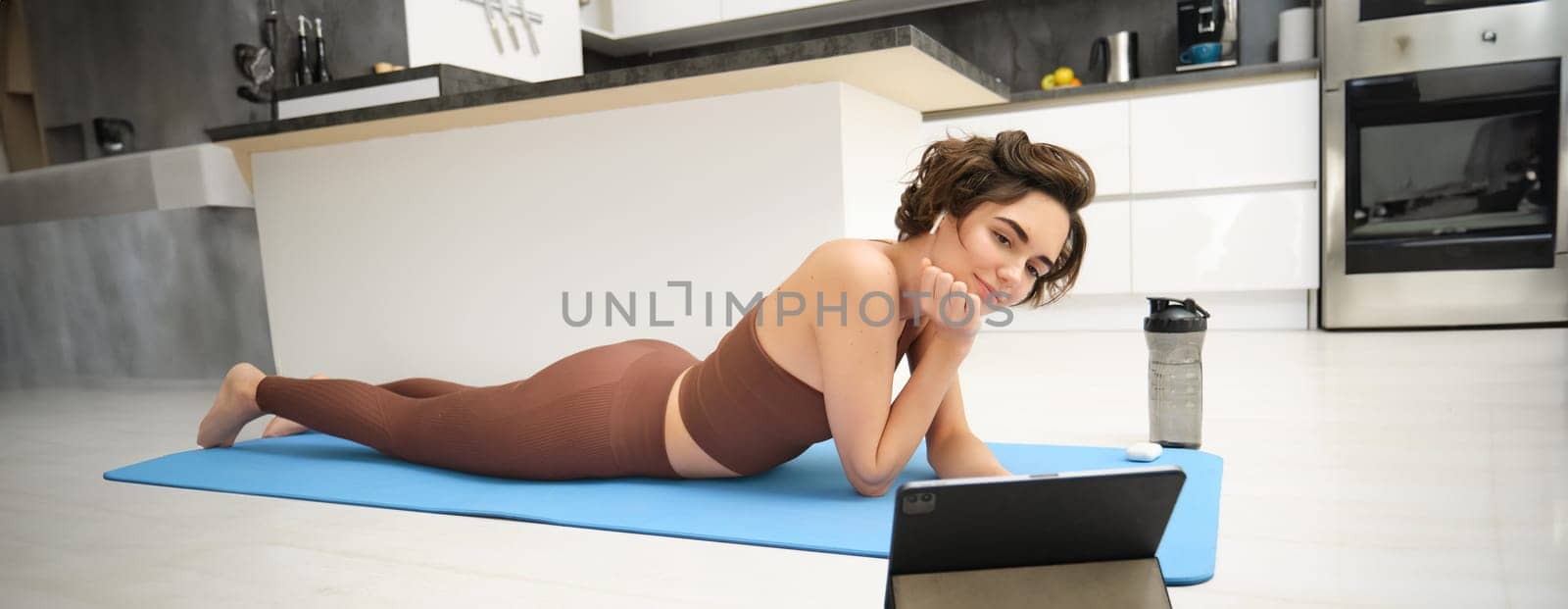 Young fitness woman, girl in tracksuit lying on fitness mat, looking at yoga, pilates video tutorial on tablet, does workout from home, uses gym app, training at home.