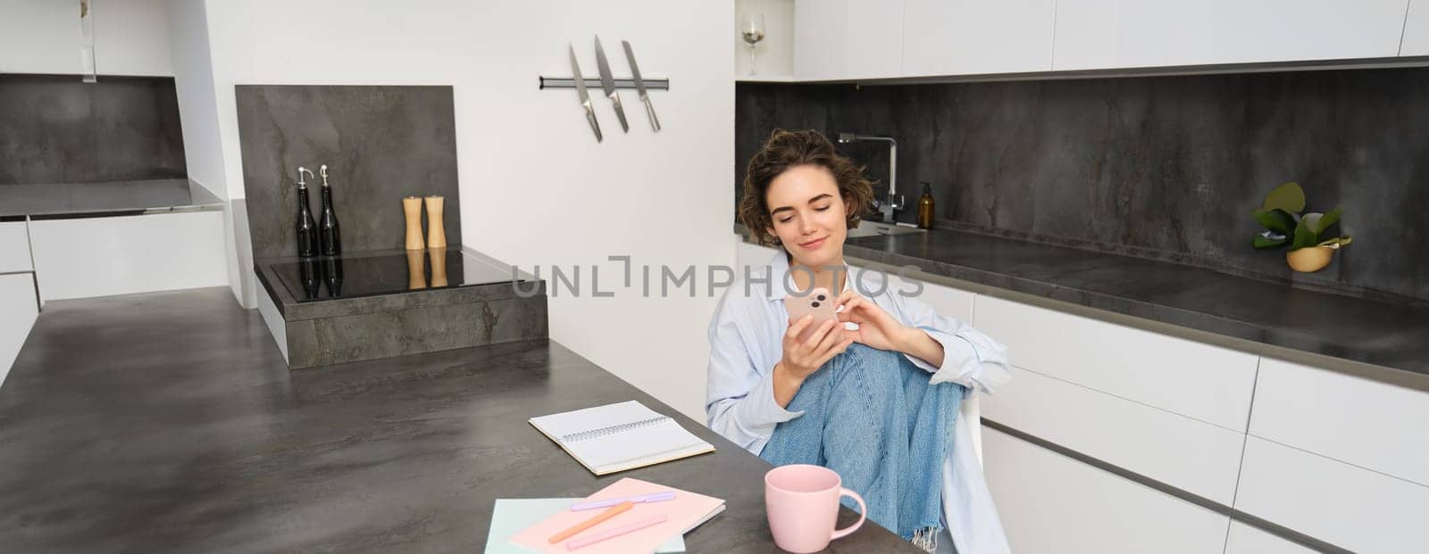 Beautiful girl with smartphone, sits at home, does homework, watches smth on mobile phone app by Benzoix