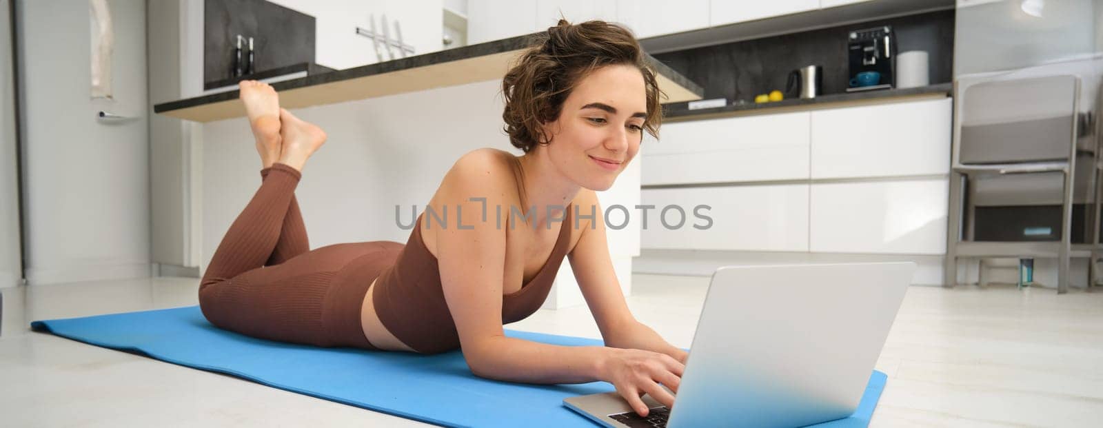 Portrait of beautiful sportswoman, fitness girl watching videos on laptop during workout, follow online pilates videos while exercise at home, wearing activewear in kitchen.