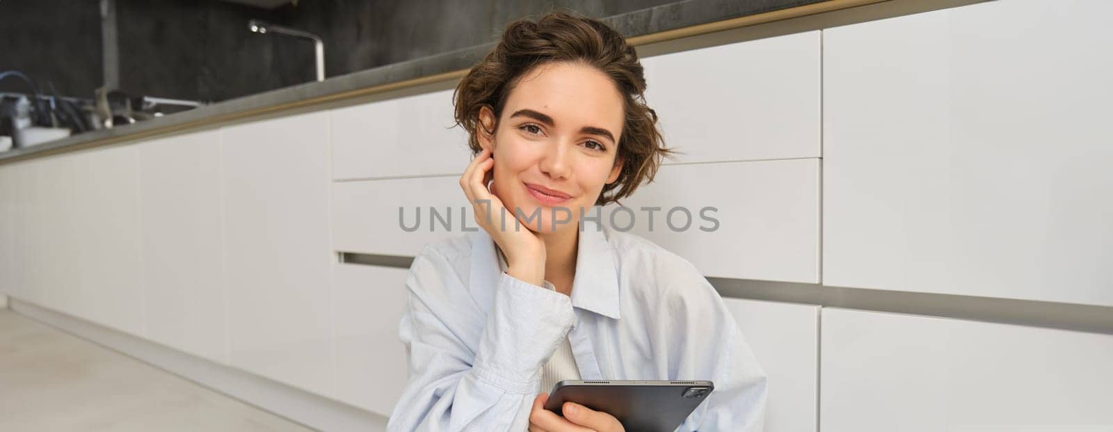 Portrait of beautiful brunette woman, holding black digital tablet, smiling and gazing gently at camera, sitting on floor at home by Benzoix