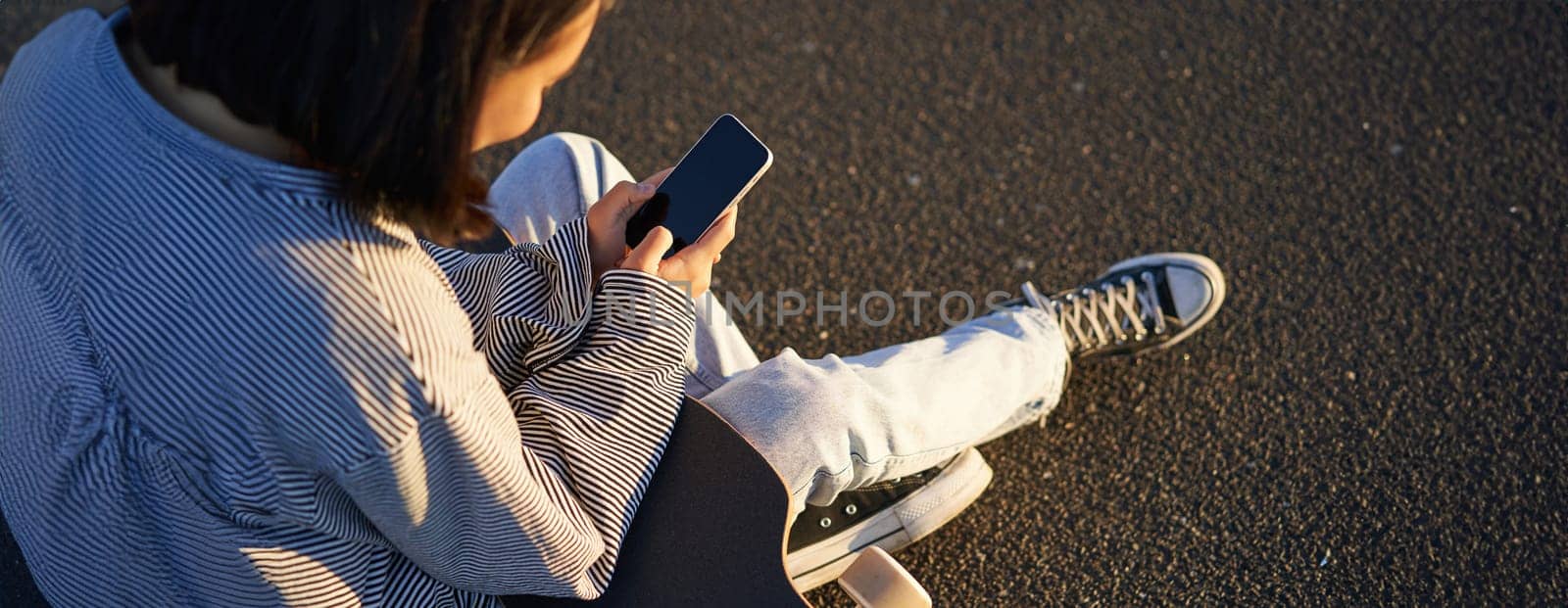 Close up of skater teen girl sits on skateboard, types message on smartphone, looks at mobile phone screen by Benzoix