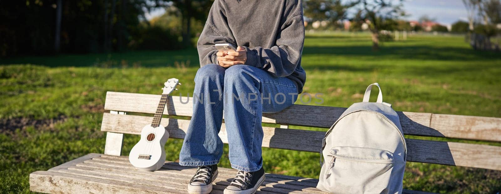 Cropped shot of teen girl body, sitting on bench with ukulele, using smartphone, hands holding mobile phone.