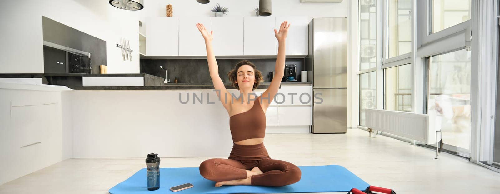 Smiling young woman in activewear, sitting on mat at home, raising arms up while meditating, practice yoga, doing mindful workout. Sport and wellbeing