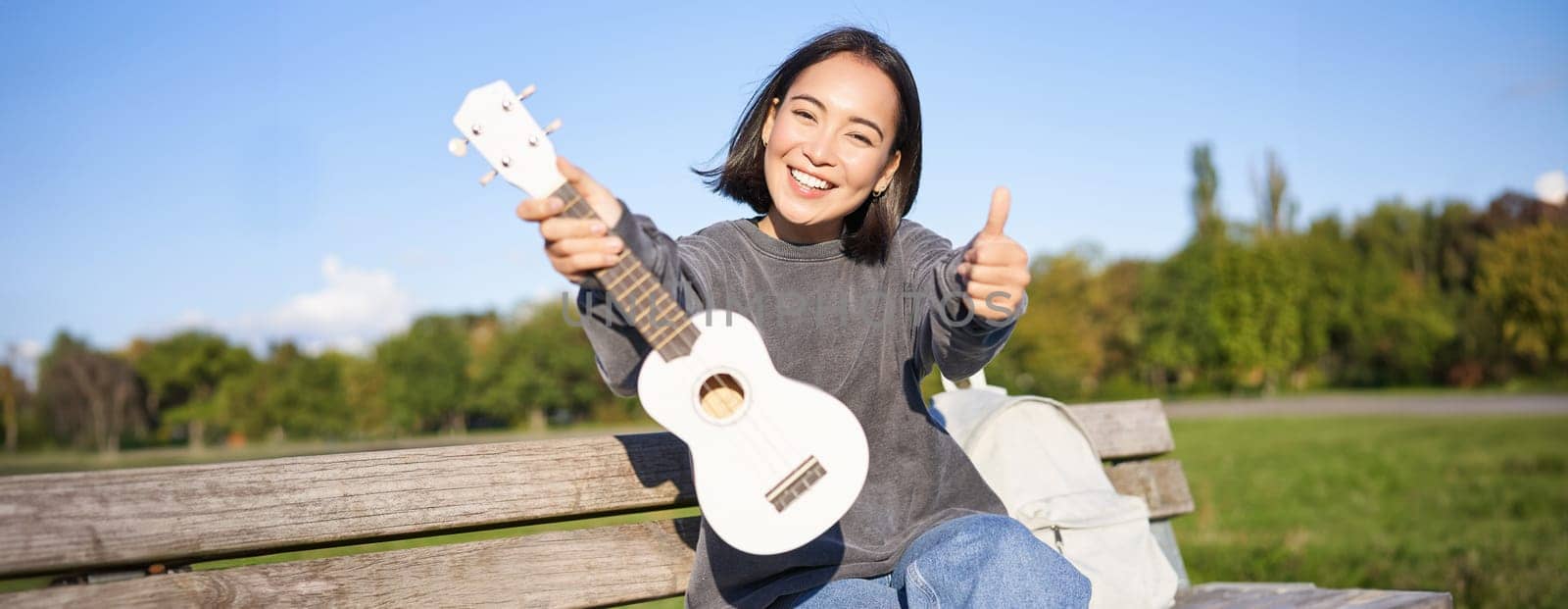 Happy asian girl shows ukulele and thumbs up, demonstrates her new musical instrument, learns how to play in park, sits on bench. Copy space