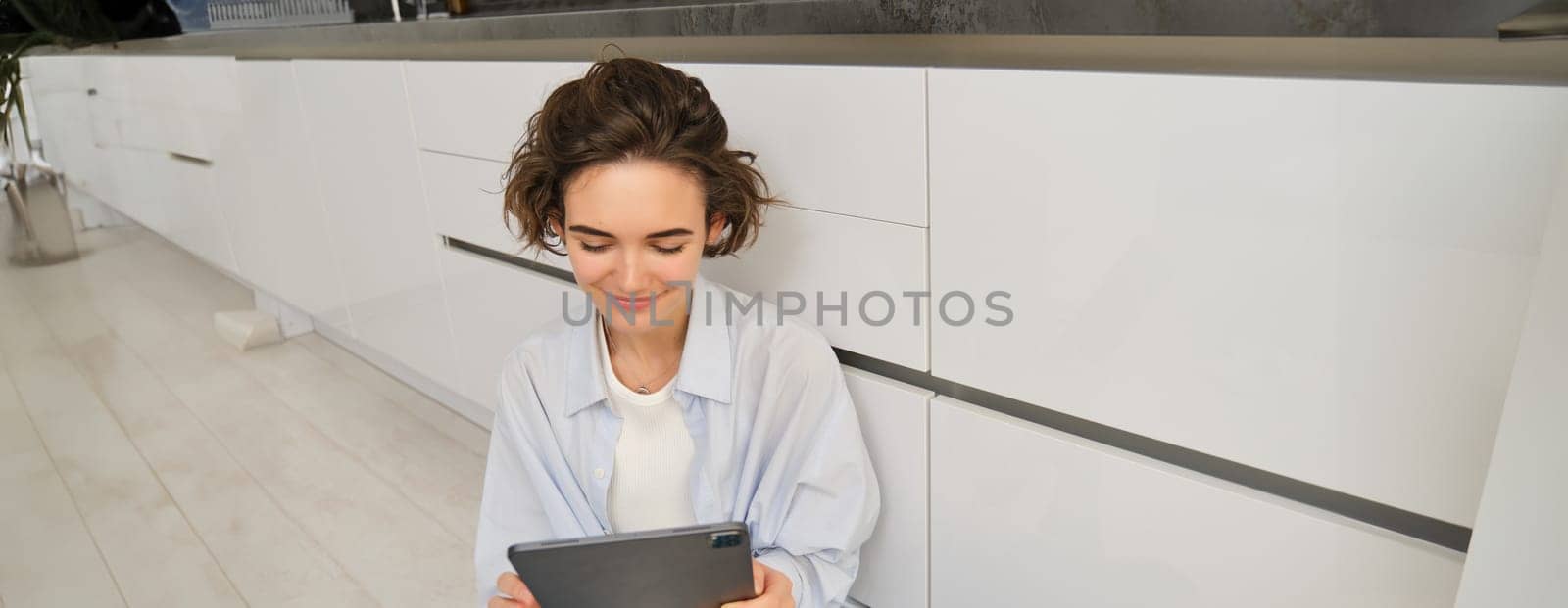 Portrait of woman looking at her tablet, reading online, watching videos on digital device, sitting on floor at home. Copy space