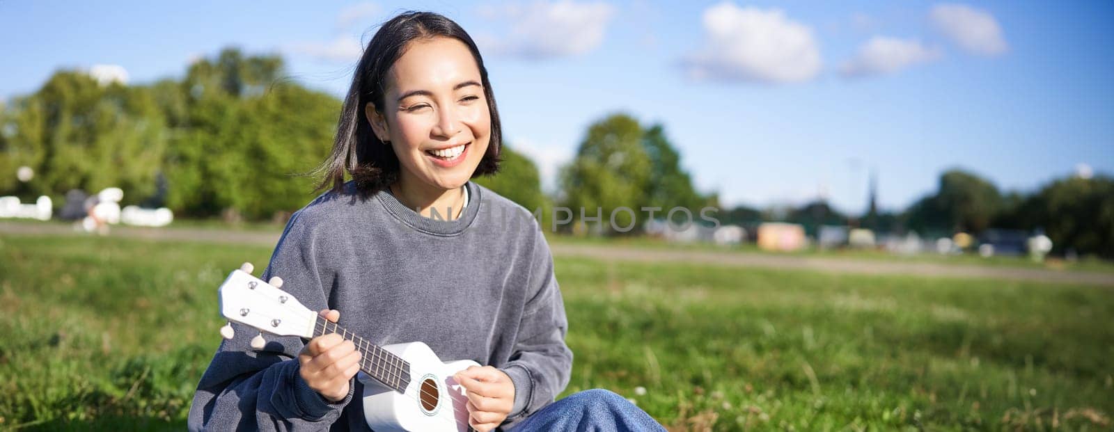 Carefree asian girl singing and playing ukulele in park, sitting on grass, musician relaxing on her free time outdoors.