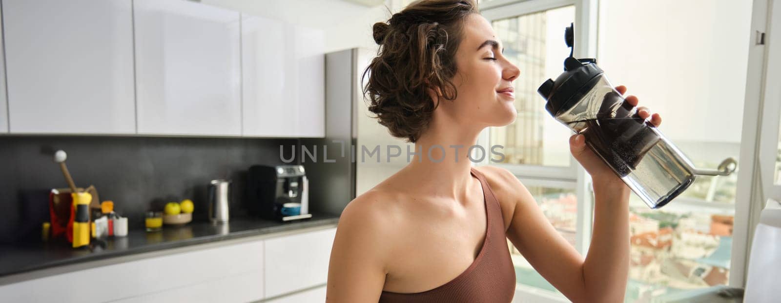 Healthy lifestyle and sport. Smiling brunette female athlete, fitness girl drinks water from gym bottle with pleased face, standing at home in kitchen.