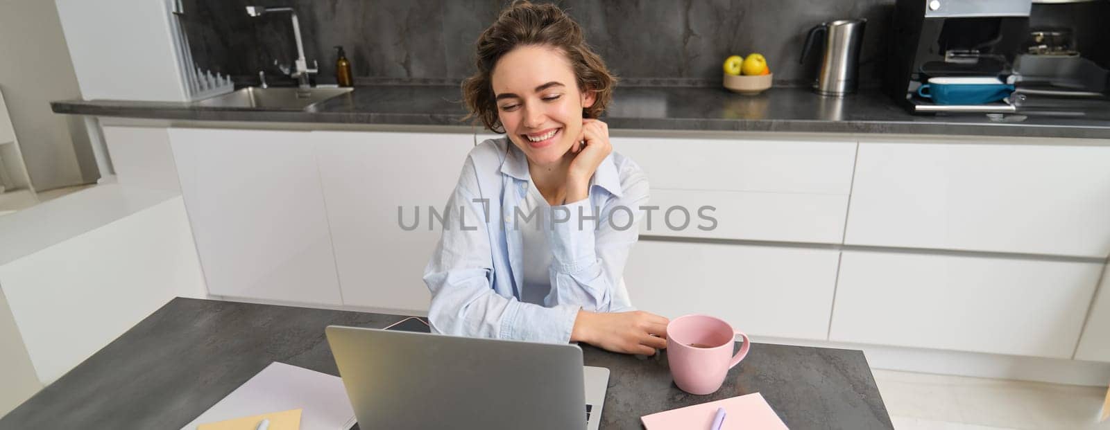 Young woman freelancer works from home, looks at laptop screen and smiles, does homework in kitchen.