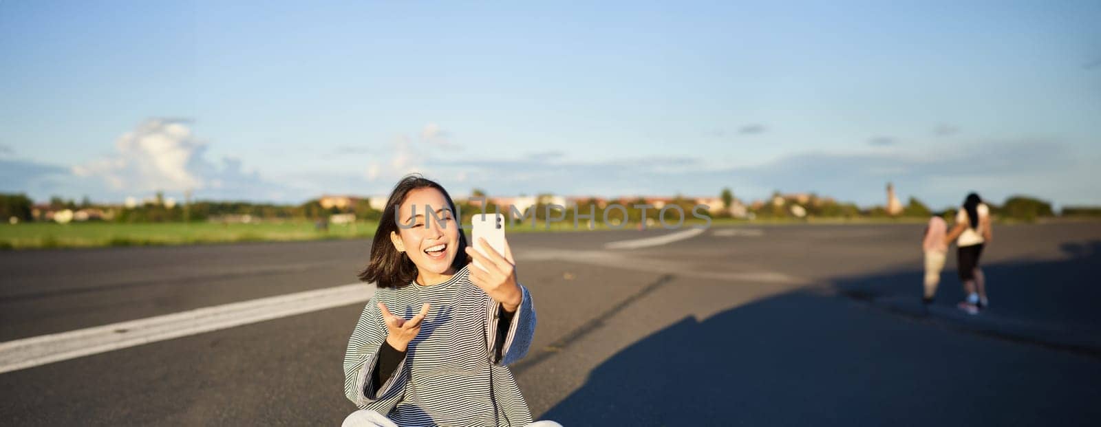 Positive teen asian girl takes selfie, video chats on mobile phone app, sits on skateboard and records vlog.