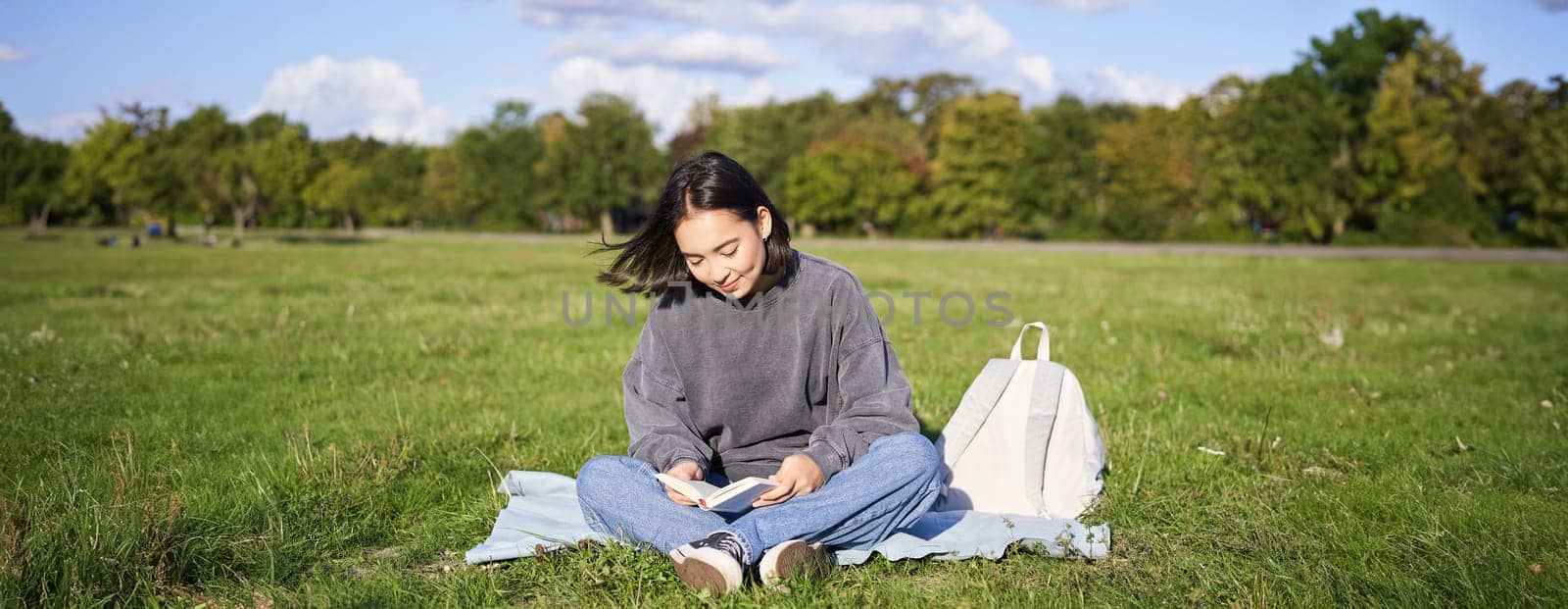 Portrait of asian girl reading book, sitting on her blanket in park, with green grass, smiling happily.