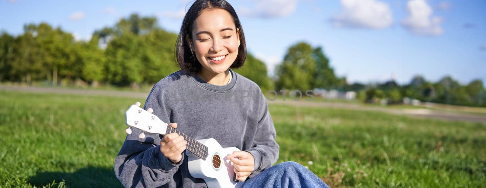 Carefree asian girl singing and playing ukulele in park, sitting on grass, musician relaxing on her free time outdoors by Benzoix
