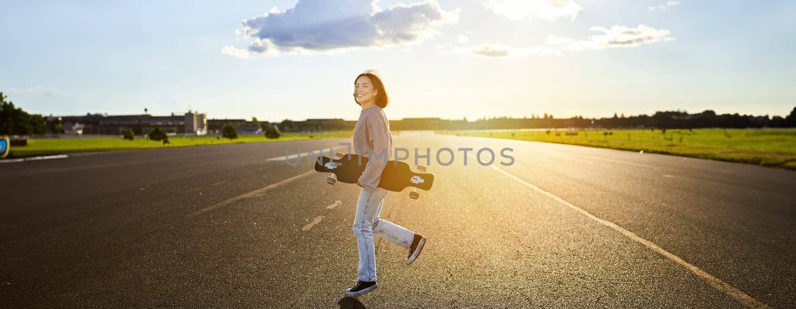 Asian girl with skateboard standing on road during sunset. Skater posing with her long board, cruiser deck during training.