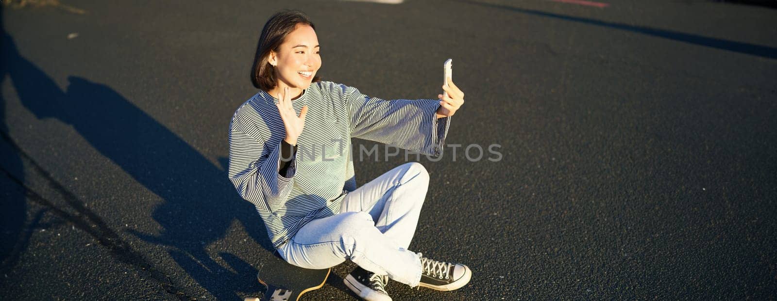Positive teen asian girl takes selfie, video chats on mobile phone app, sits on skateboard and records vlog.