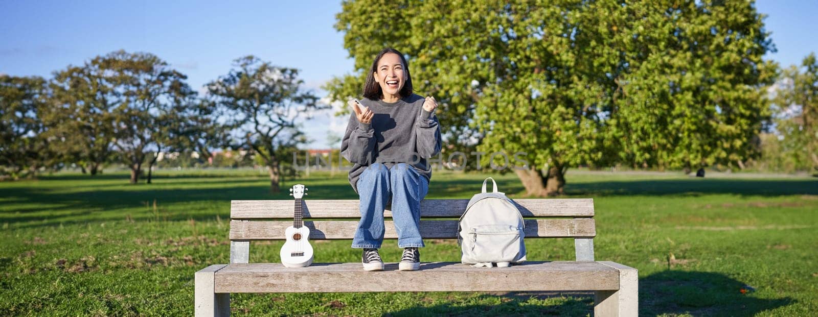 Excited girl looks at smartphone and celebrates, wins on mobile phone, sits with ukulele and backpack in park on bench on sunny day by Benzoix