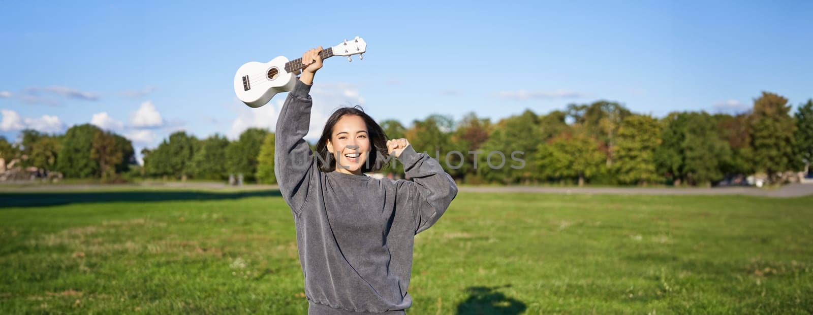 Upbeat young woman dancing with her musical instrument. Girl raises her ukulele up and pose in park on green field by Benzoix