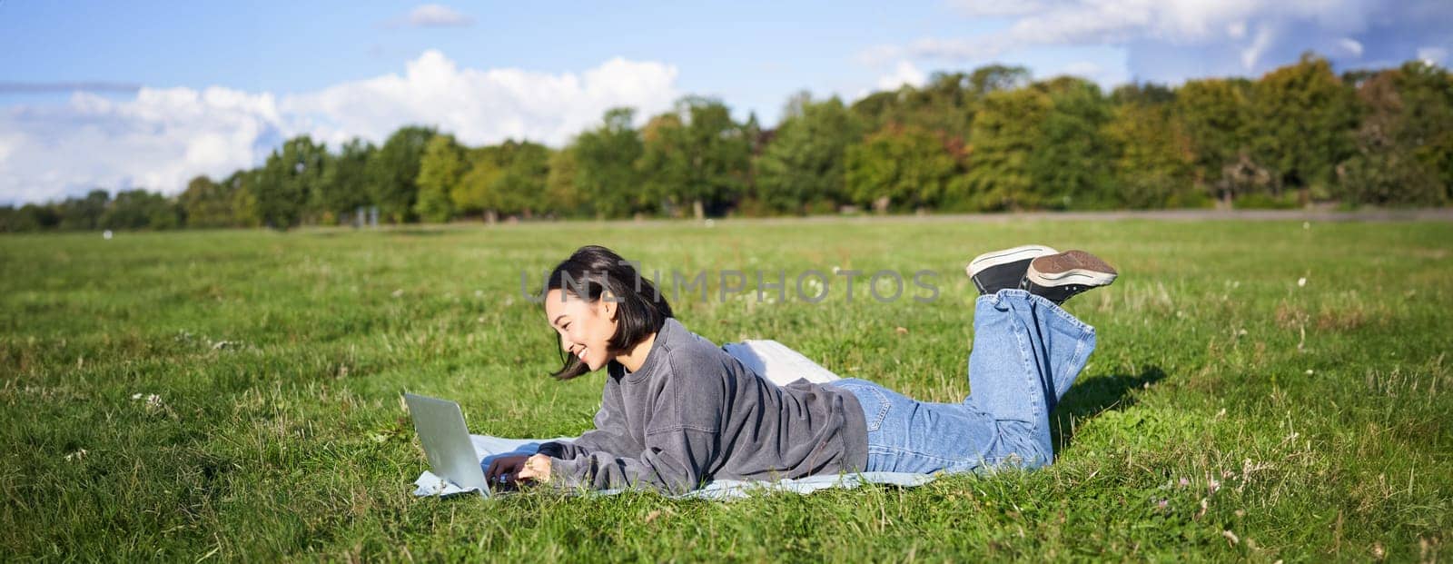 Portrait of young happy asian girl lying on blanket in park, watching videos and browsing internet on her laptop.