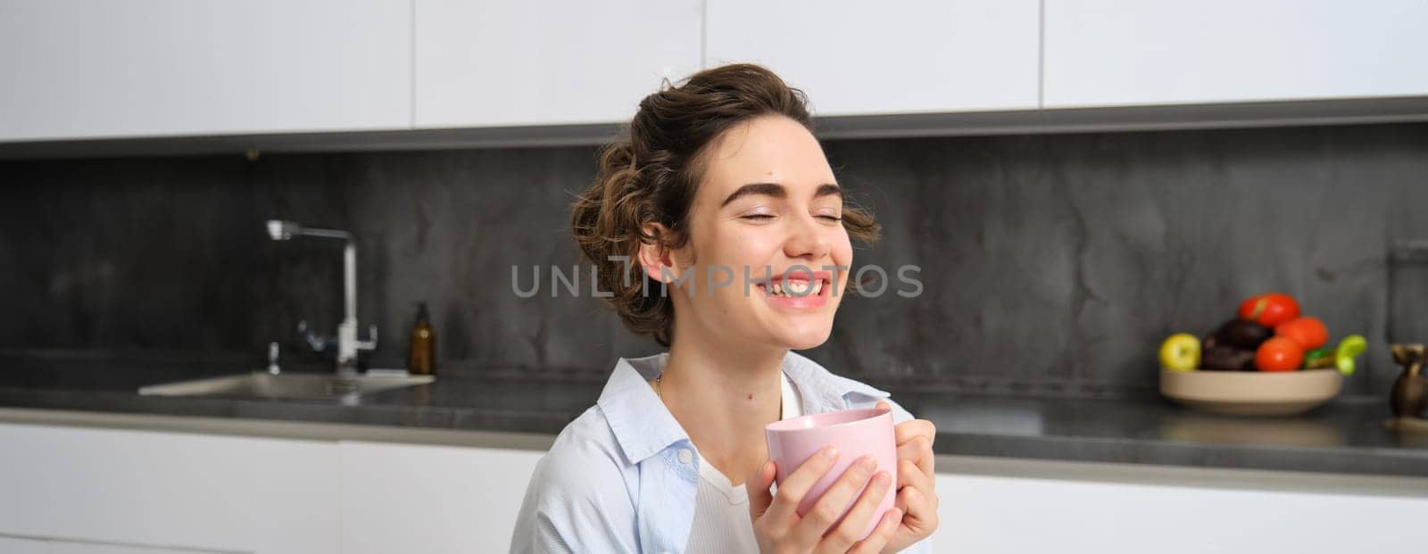 Happy brunette woman drinking coffee at home. Girl with pink cup, sits in her kitchen and enjoys relaxing morning weekend.