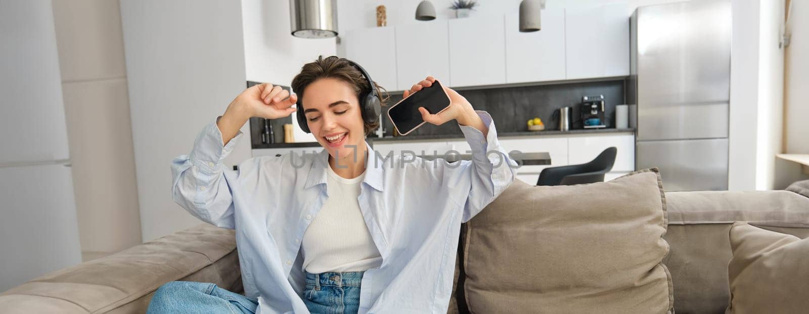 Portrait of beautiful, happy young woman, listening music in headphones, holding smartphone and dancing on sofa at home, enjoys spending time indoors alone by Benzoix