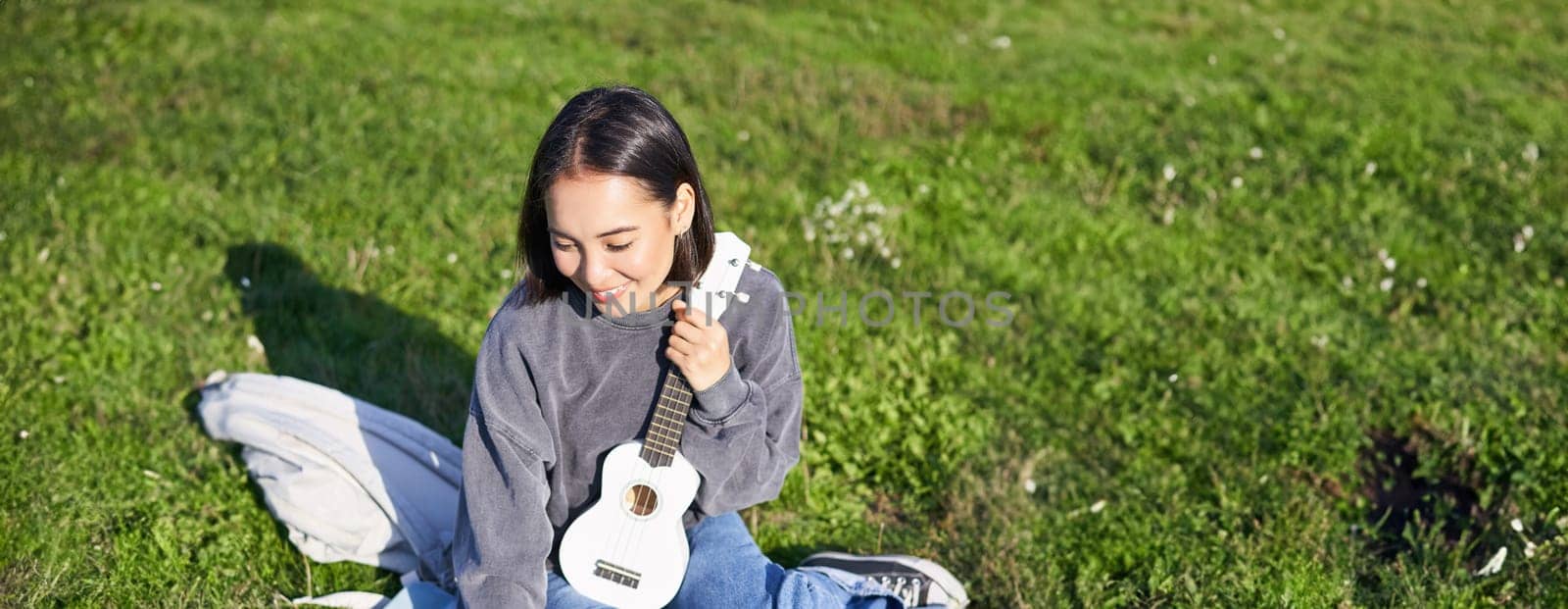 Smiling asian girl learns how to play ukulele on laptop, video chat with music teacher, sitting with instrument in park on grass.