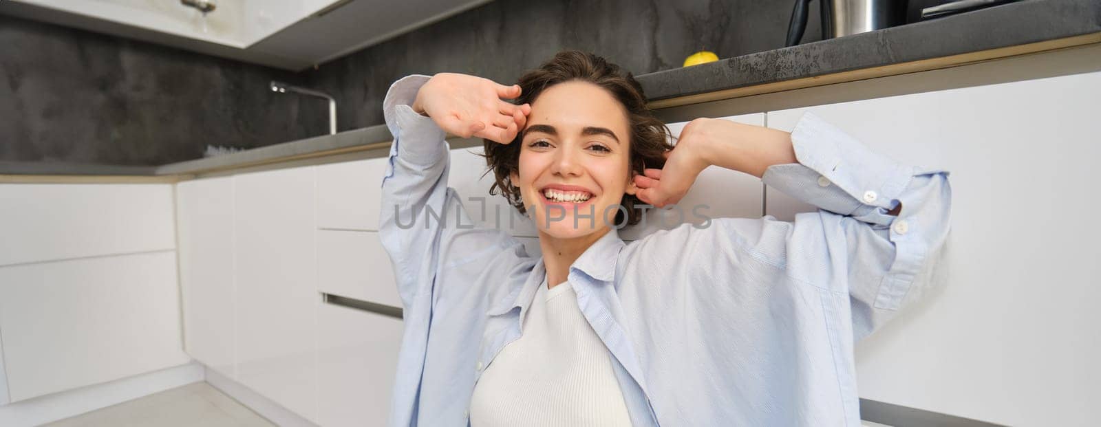 Close up portrait of beautiful smiling woman, stretching hands with pleased face, feels tired, sits on kitchen floor at her home.