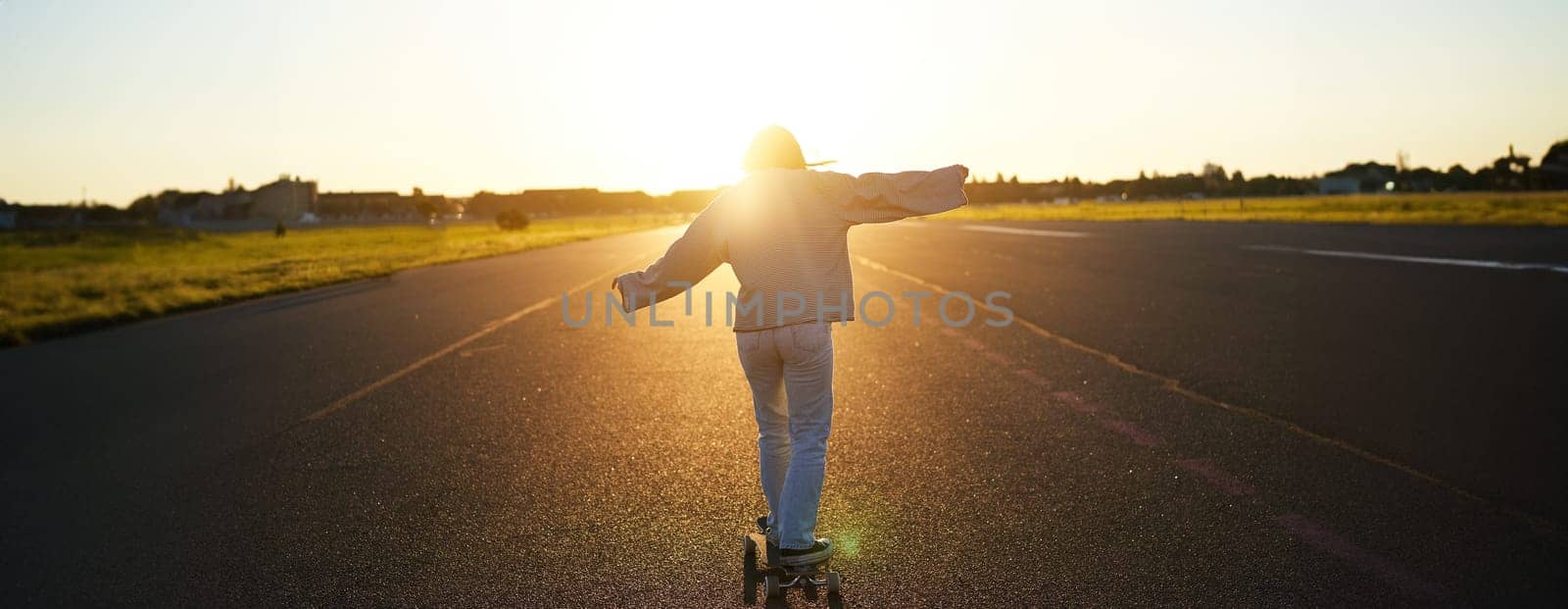 Rear view photo of young girl riding skateboard towards sunlight. Happy young woman on her cruiser, skating on longboard by Benzoix