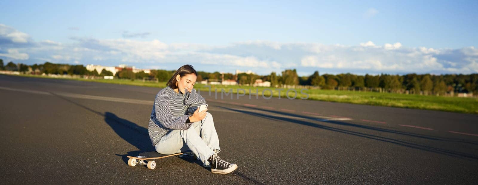 Portrait of young korean girl sitting on her skateboard on road, looking at smartphone, chatting on mobile app.