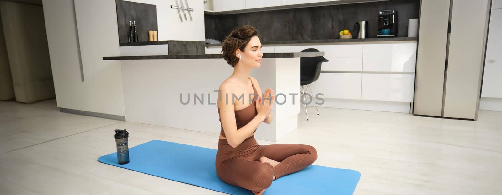 Portrait of fitness girl in kitchen, sits on yoga mat, meditates, listens meditation music in headphones, uses workout app on smartphone, does exercises at home.
