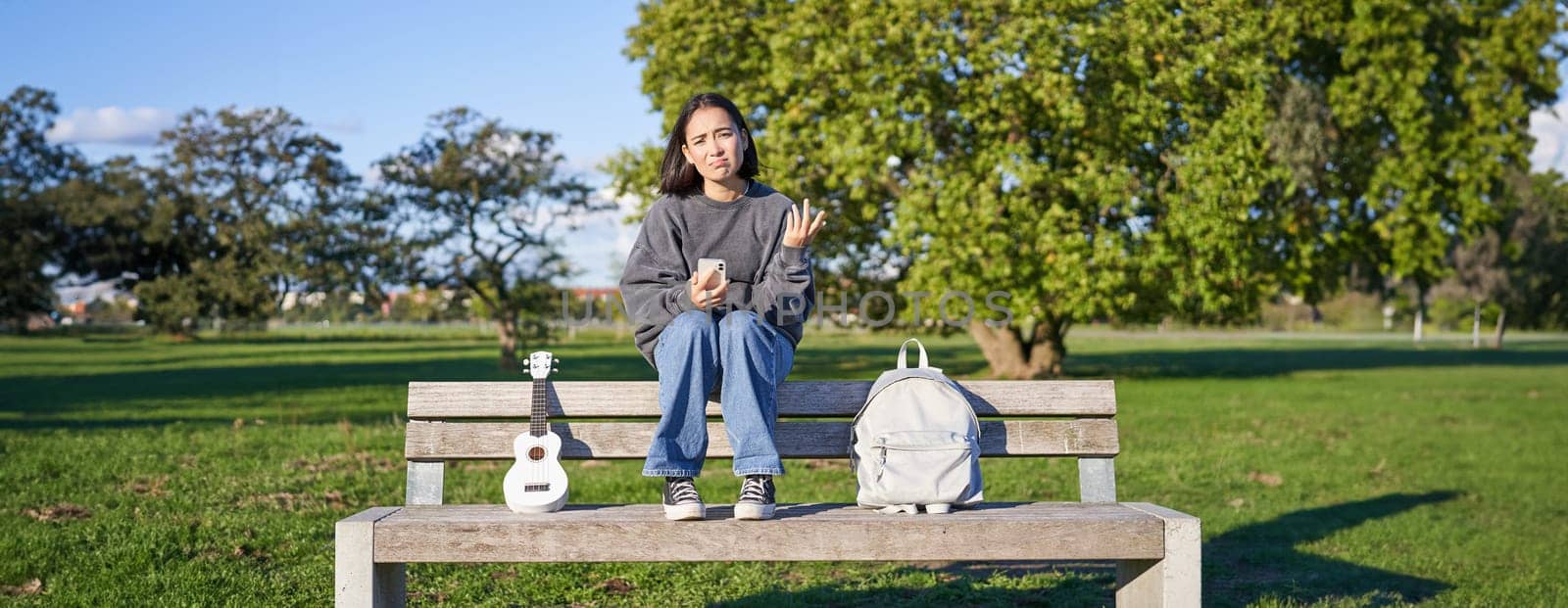 Girl with sad face, sitting on bench with smartphone and ukulele, looking upset and disappointed, being alone in park.