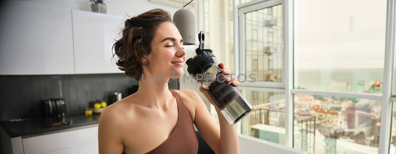 Sport and fitness concept. Portrait of young woman athlete drinking water from gym bottle, smiling with pleased face, wearing acrivewear, standing in kitchen.