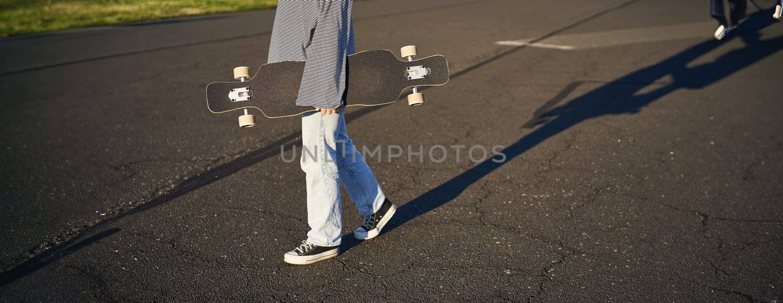 Cropped shot of teen girl body, holding cruiser longboard in hand, walking in sneakers on road in jeans and sweatshirt. Young woman skater with skateboard.