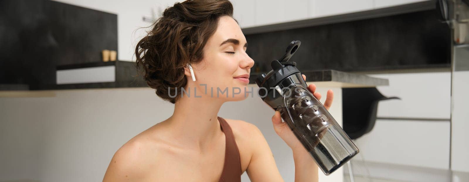 Smiling active young woman in sportswear, sits on yoga mat with water bottle, drinks stays hydrated during workout training session from home, fitness in the house on floor.