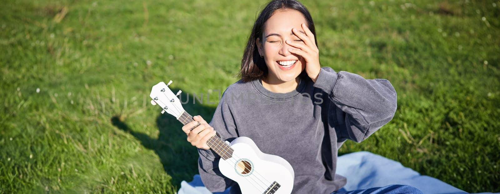 Smiling asian girl with ukulele, playing in park and singing, lifestyle concept. Copy space