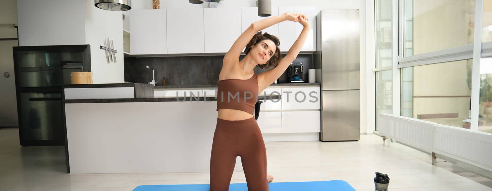 Home training. Young woman workout at home, stretching her body, doing fitness exercises on yoga rubber mat, listening to gym app instructions on smartphone.