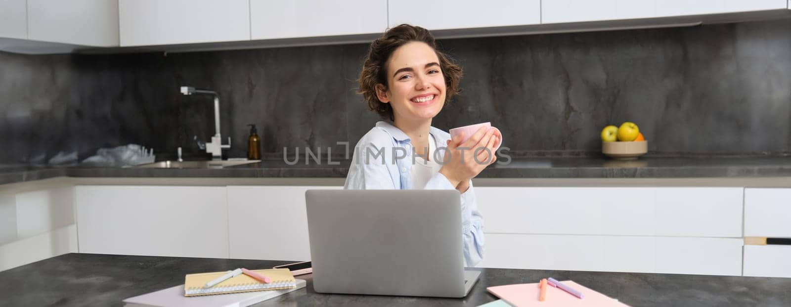 Smiling brunette girl sits in kitchen with laptop, drinks coffee and works from home.