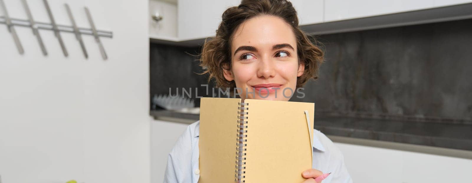 Portrait of smiling brunette girl, holds her planner, writes in notebook and looks happy, laughs, does homework, makes notes, sits at home in kitchen.