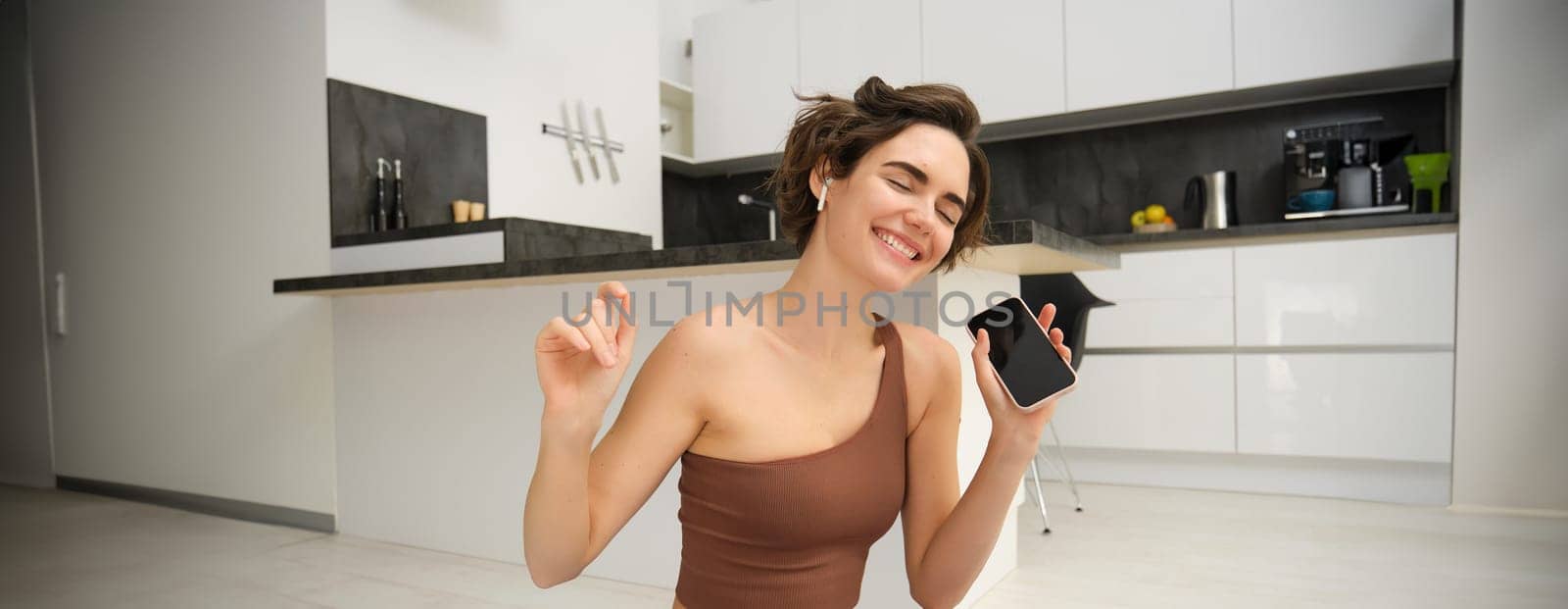 Active lifestyle and health. Happy young woman does workout, sits and dances with smartphone in hands, listens music in wireless earphones during fitness training session, yoga on floor mat.