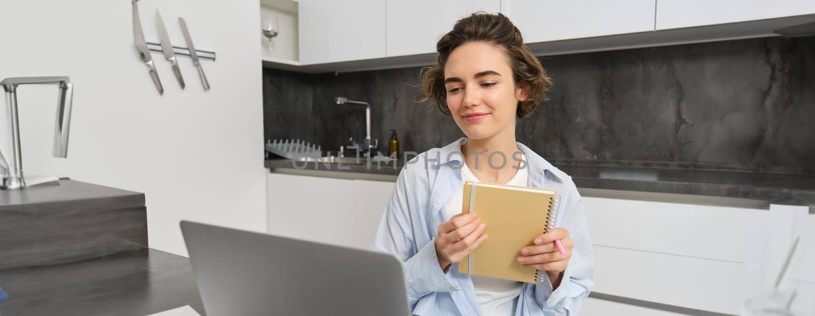 Portrait of businesswoman works from home, looks at laptop and writes down information, makes notes, studies online on course website.