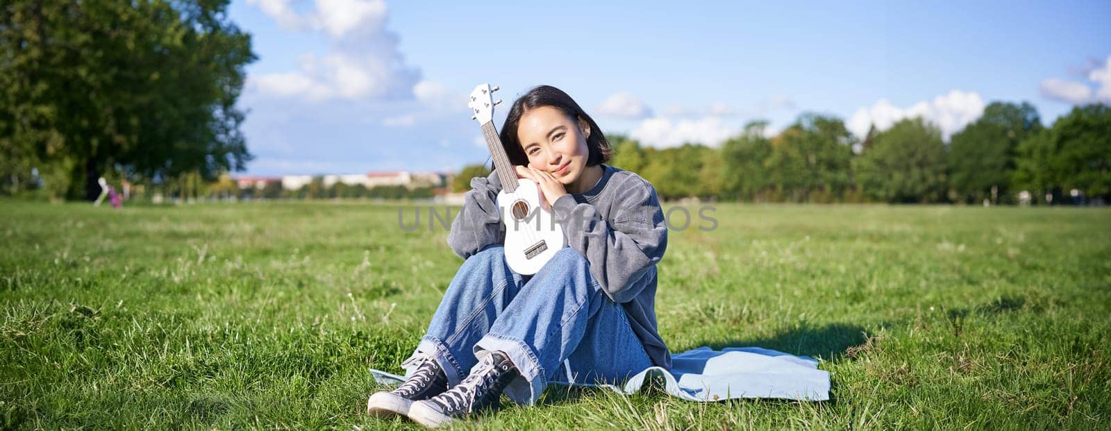 Cute romantic asian girl sitting with her instrument in park, hugging ukulele and smiling, resting outdoors.