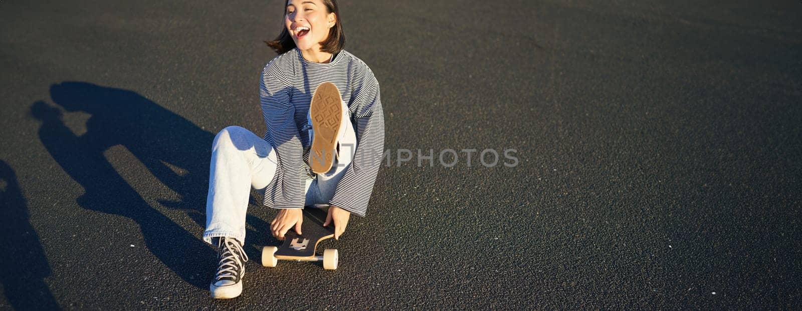 Portrait of beautiful asian girl skating, sitting on her skateboard and smiling. Cute teenager with longboard, skating on road by Benzoix