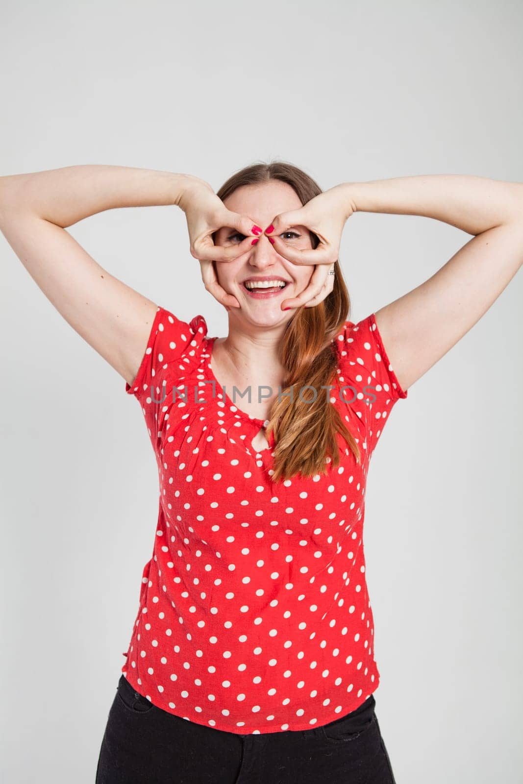 Studio portrait of smiling attractive woman wearing spotty red blouse looking through finger goggles