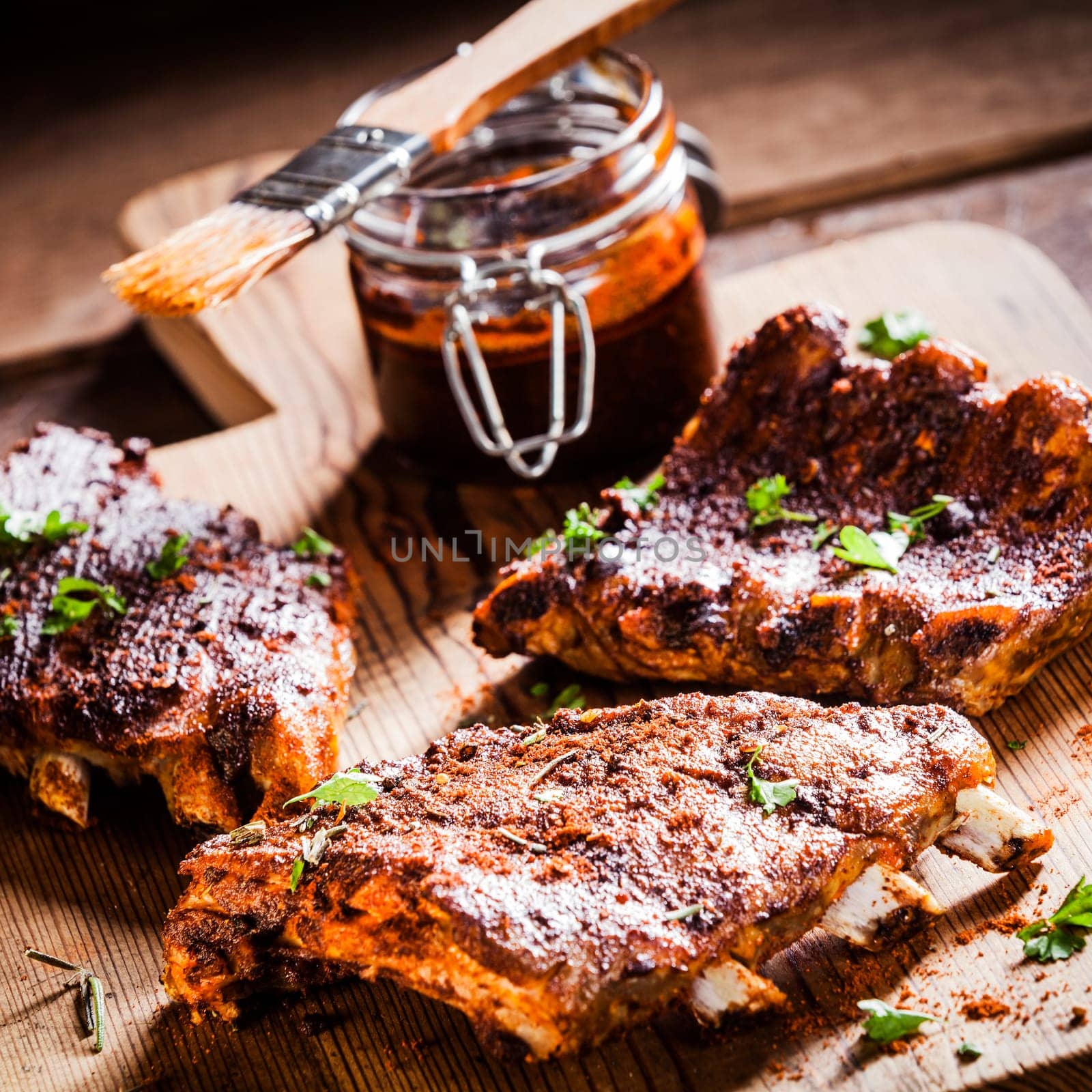Three portions of barbecued ribs in a spicy marinade seasoned with pepper and fresh herbs being prepared in a country kitchen with a jar of basting sauce