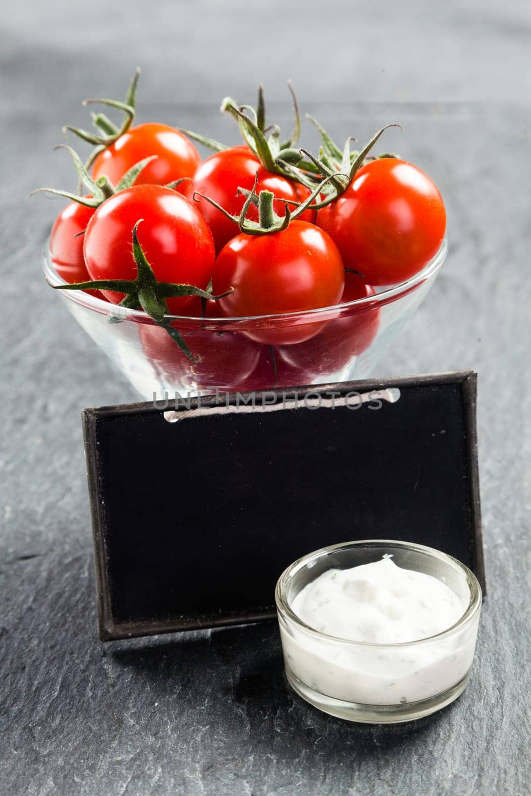 Glass bowl of fresh ripe red cherry tomatoes and savory creamy vinaigrette sauce on a slate background with a blank vintage school slate with copyspace for your text