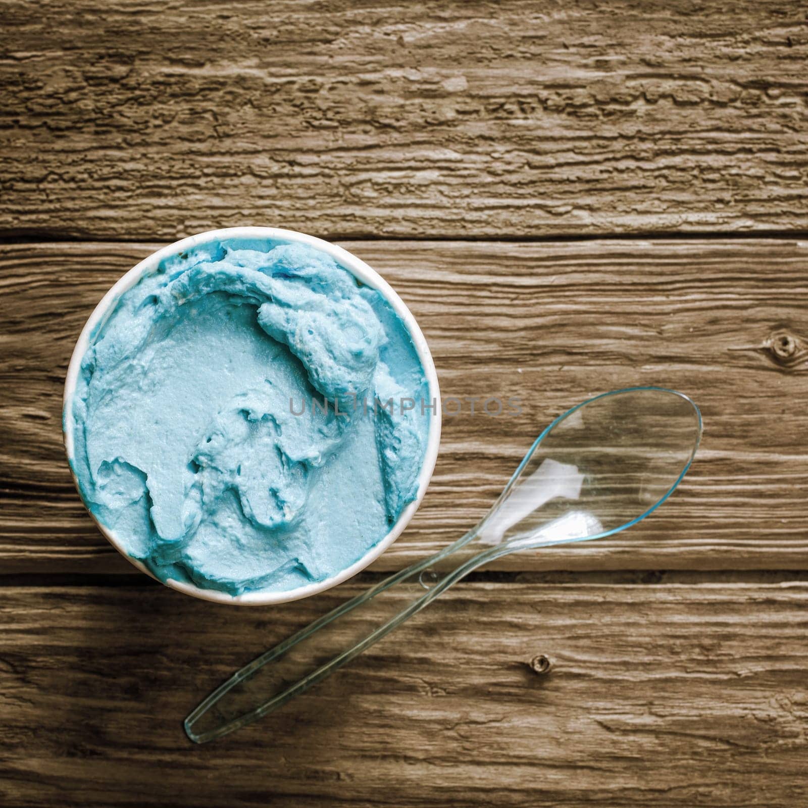 Close up overhead view of a tub of gourmet blue Italian ice cream with a plastic spoon served on an old wooden table, in square format