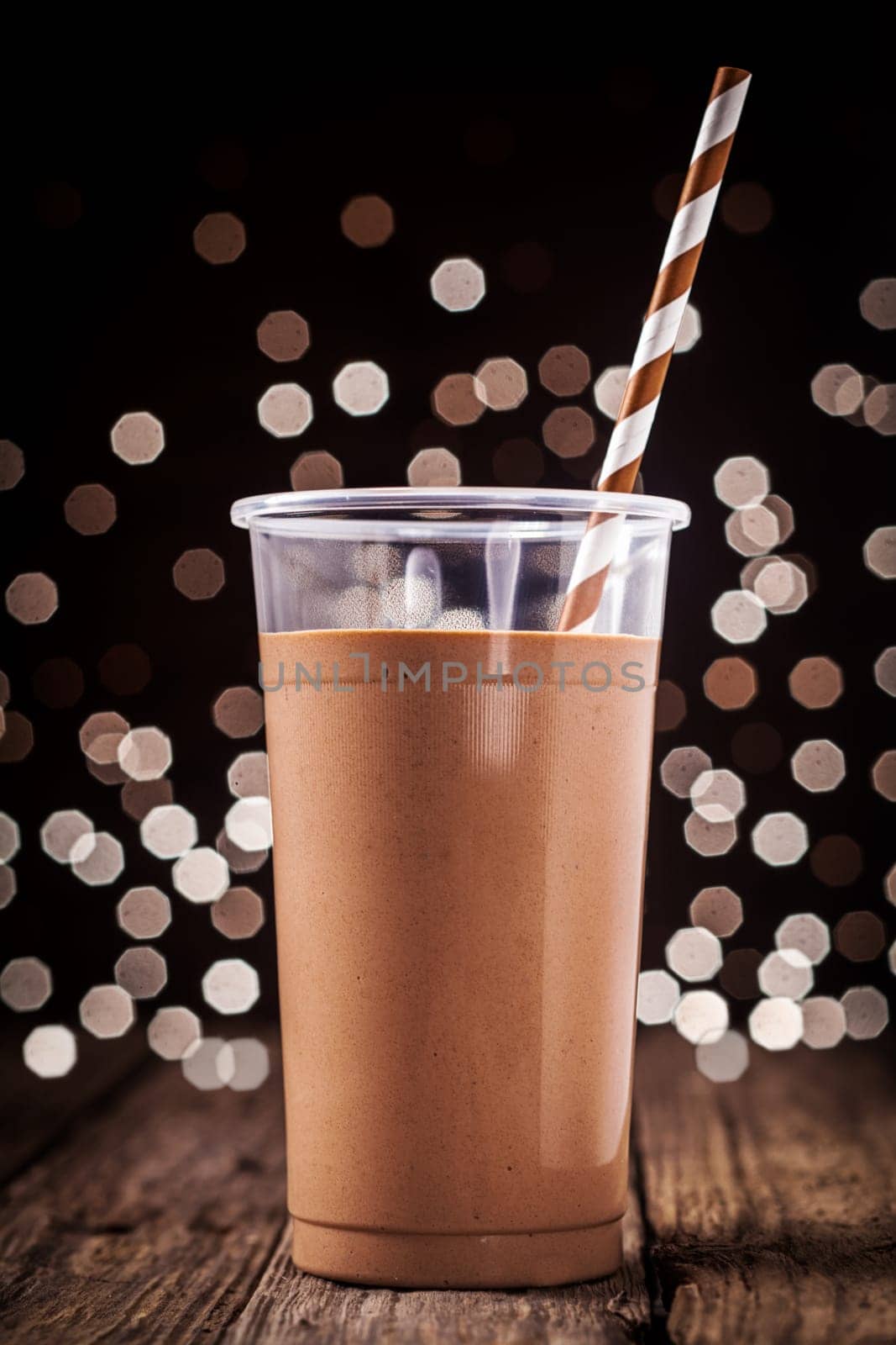 Plastic glass of delicious creamy chocolate smoothie or milkshake against a background of sparkling festive party lights