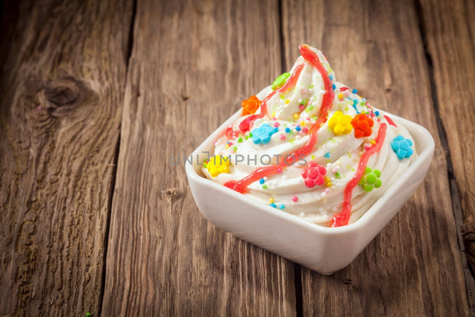 Colorful party ice cream dessert frozen yogurt with a swirl of vanilla ice cram topped with bright multicolored candy and sprinkles drizzled with syrup on an old textured wooden table with copyspace